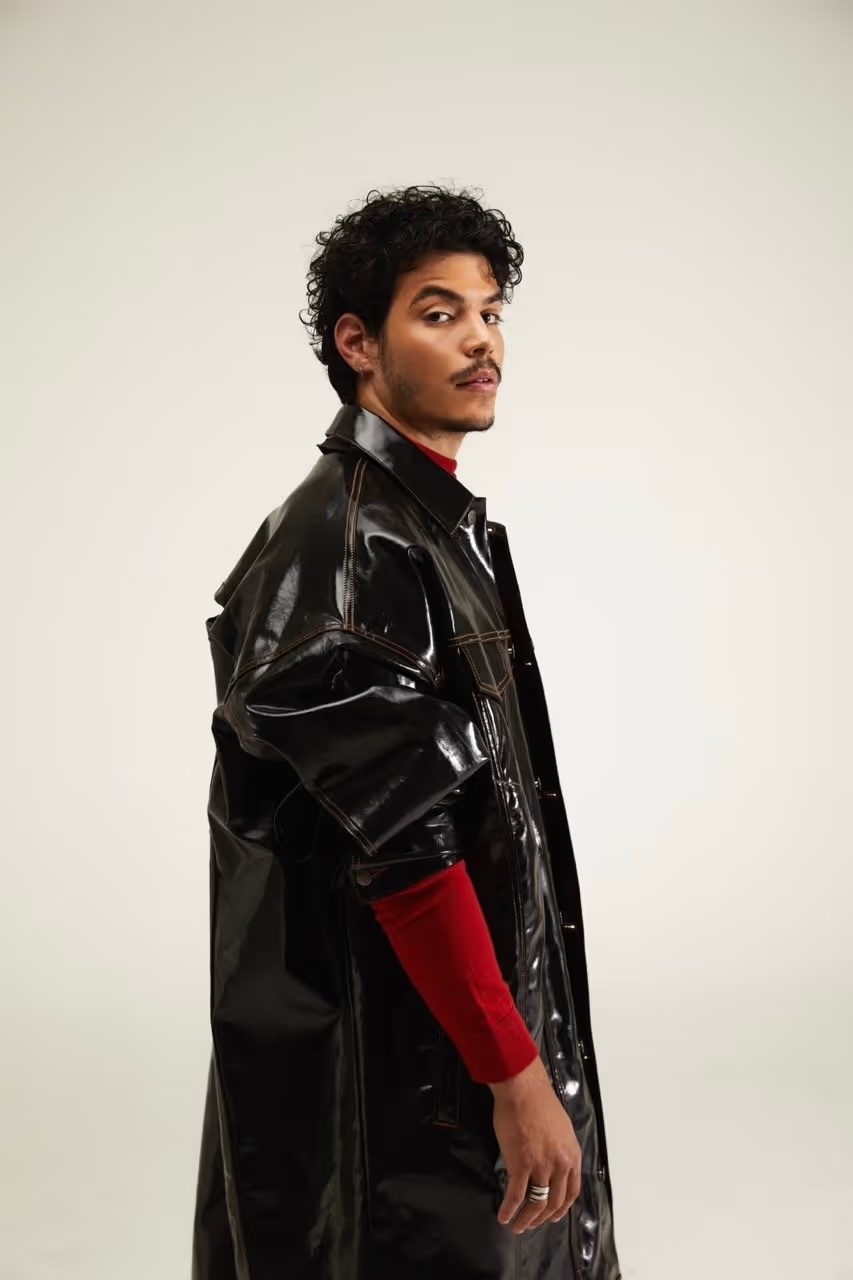 Studio shot of adult male with curly hair, wearing black vinyl trench coat over a red turtleneck against a plain white background.