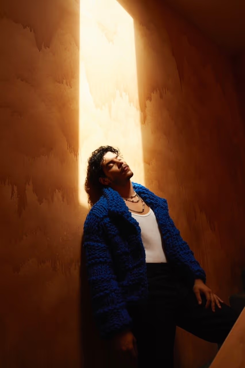 Adult male poses in an indoor space, illuminated by a bright skylight, wearing a blue textured jacket, black pants, and a white tank top.