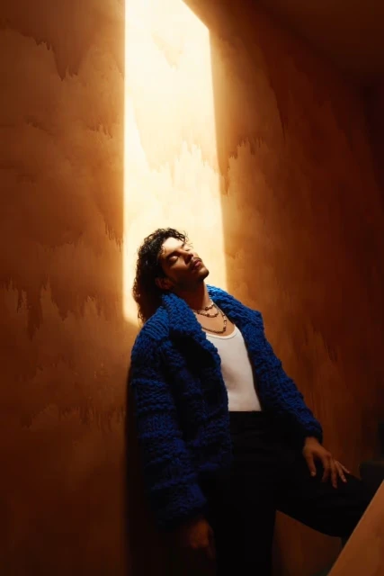 Adult male poses in an indoor space, illuminated by a bright skylight, wearing a blue textured jacket, black pants, and a white tank top.