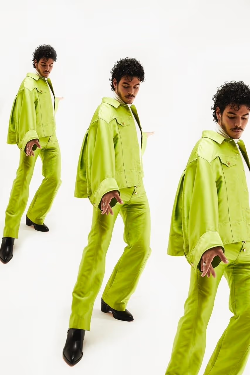 An adult male poses in a lime-green suit and black boots in a studio setting with a white background, creating a fashion editorial image.