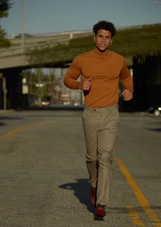 Athletic man in a brown turtleneck and trousers jogging on a city street under a bridge on a sunny day with clear blue sky.