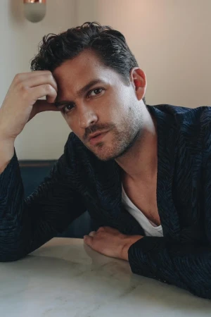 Portrait of a man in a dark patterned blazer and white undershirt resting his hand on his head at a table.