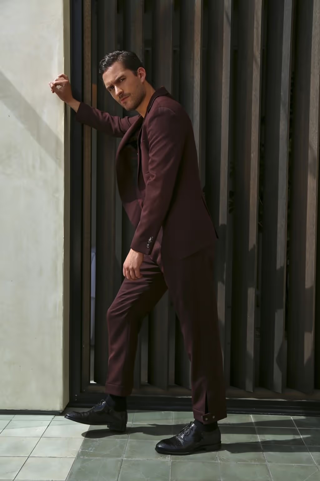 A stylish man poses confidently in a maroon suit with his hand against a wall, wearing black dress shoes and set against a dark slat wall.