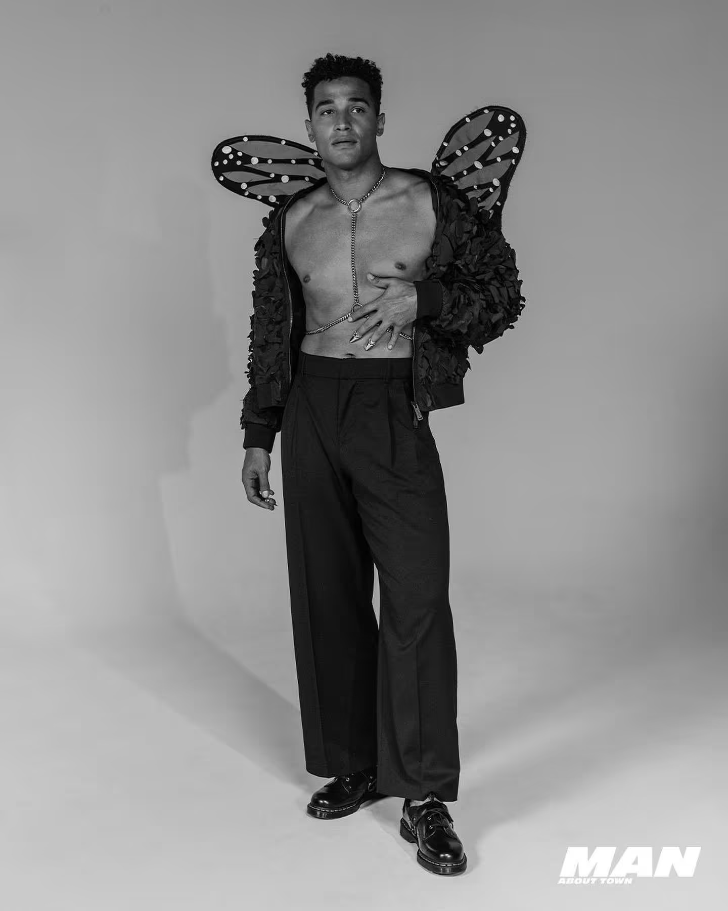 Black and white portrait of a male model wearing butterfly wings and a bomber jacket posing in front of a neutral backdrop.