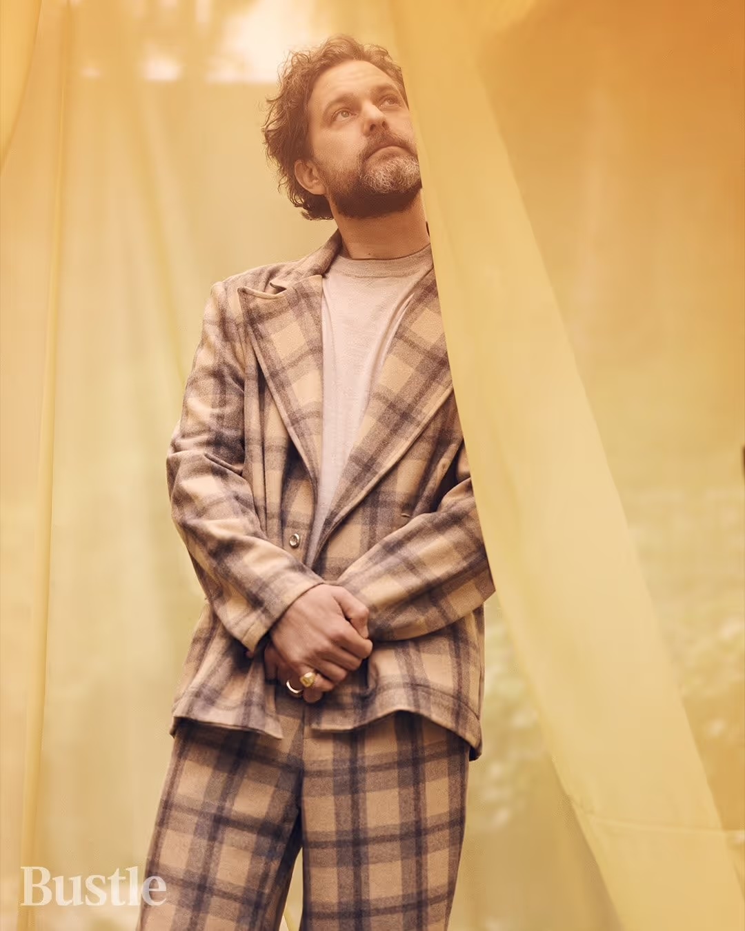 Bearded adult male in a plaid suit standing indoors by a sheer yellow curtain looking upward, posing in a three-quarter portrait shot.