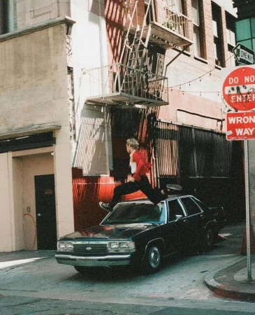 A young adult jumps across a black sedan parked on a street next to a brick building with visible fire escape and street signs.