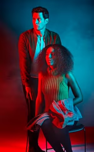 A woman sits on a stool in front of a standing man, both lit with red and blue gel lighting in a studio setting, posing for a portrait.