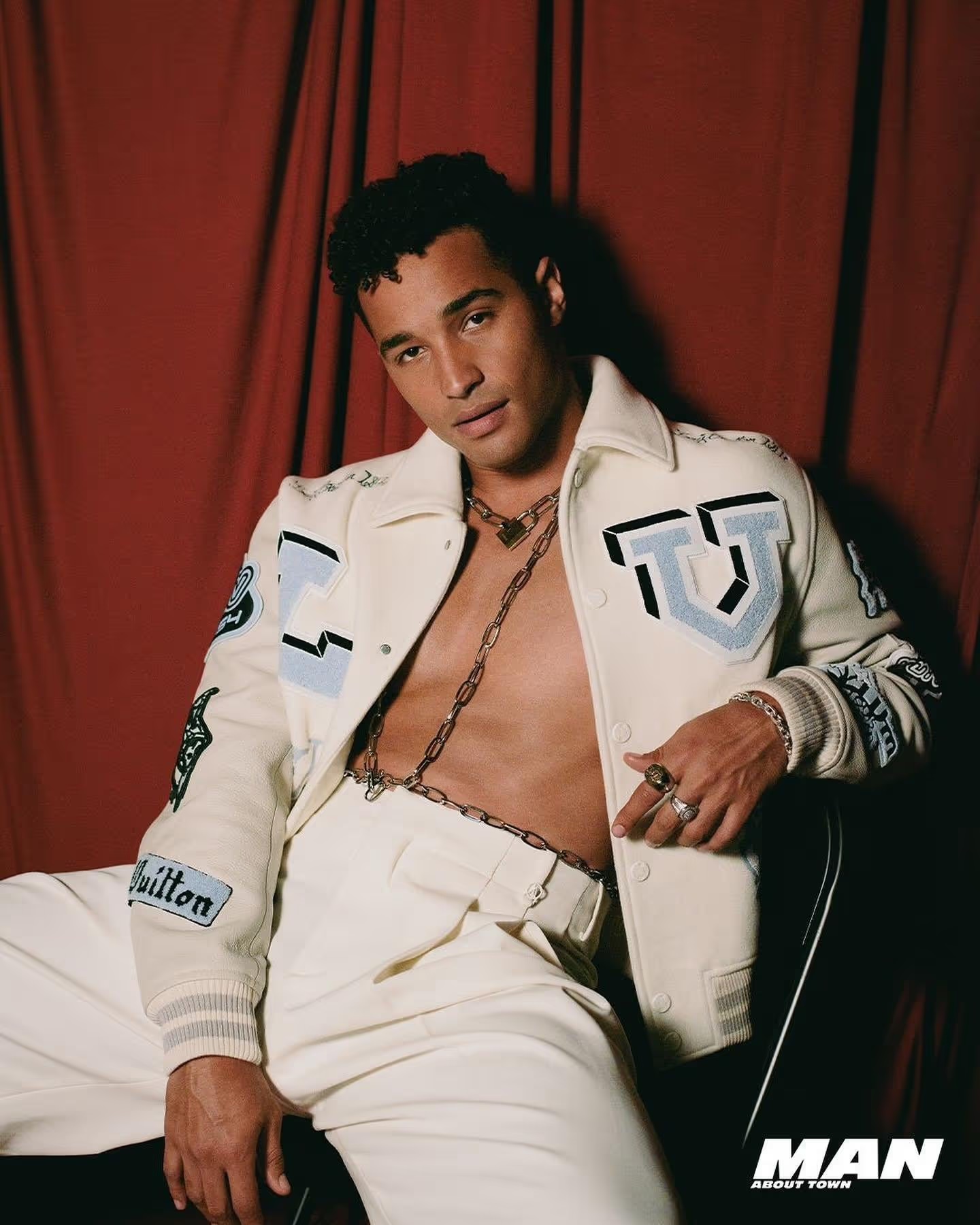 Man poses in off-white pants and jacket with patches against a red curtain. He has a chain around his neck and open jacket.