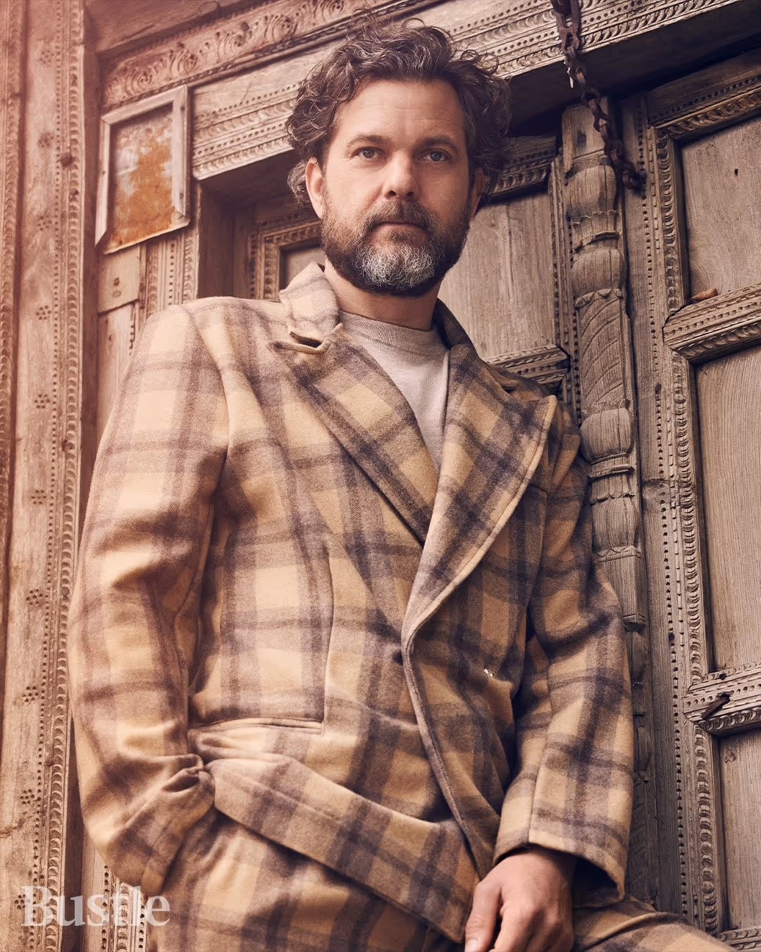Portrait of a male model in a brown plaid suit leaning against an intricately carved wooden door in soft, neutral lighting.