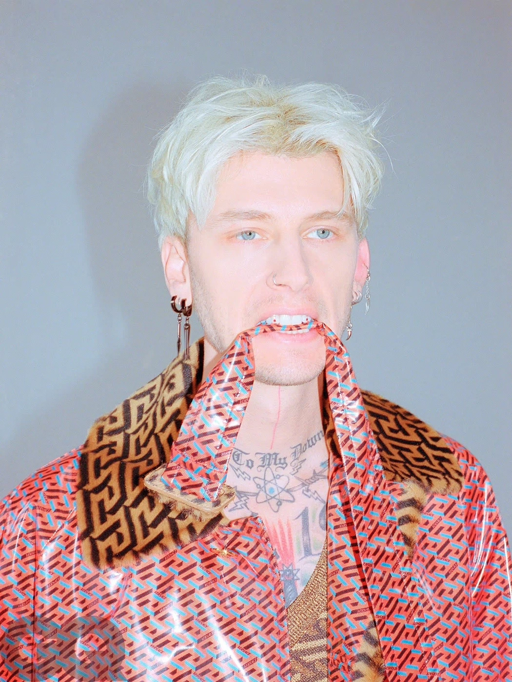 Machine Gun Kelly with dyed blonde hair biting a strap of a patterned red vinyl jacket against grey background.