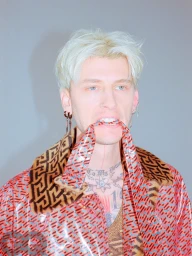 Machine Gun Kelly with dyed blonde hair biting a strap of a patterned red vinyl jacket against grey background.