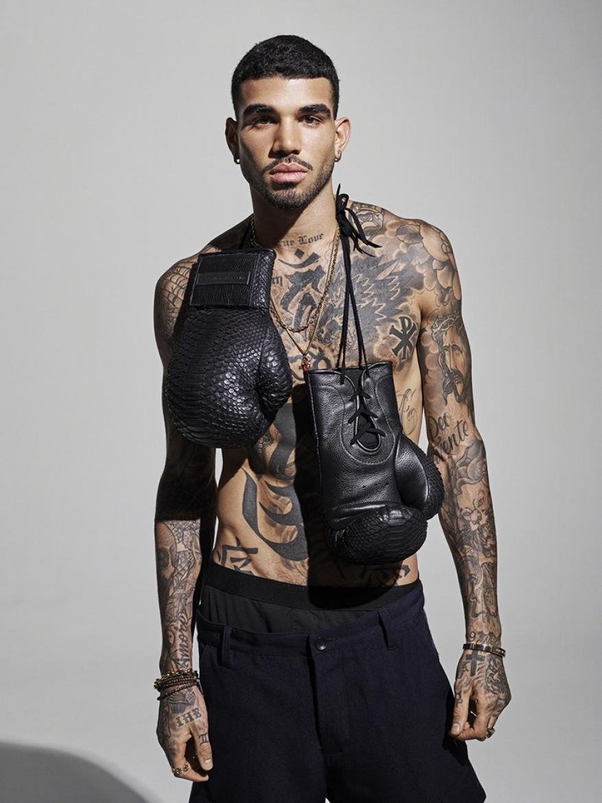 Portrait of a shirtless, tattooed man with black boxing gloves hanging around his neck in a studio with neutral lighting.