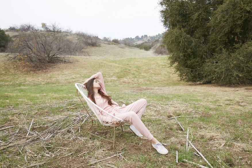 Woman in a pink sweatsuit reclines on a chair in a grassy field, surrounded by sparse trees and foliage, eyes closed.