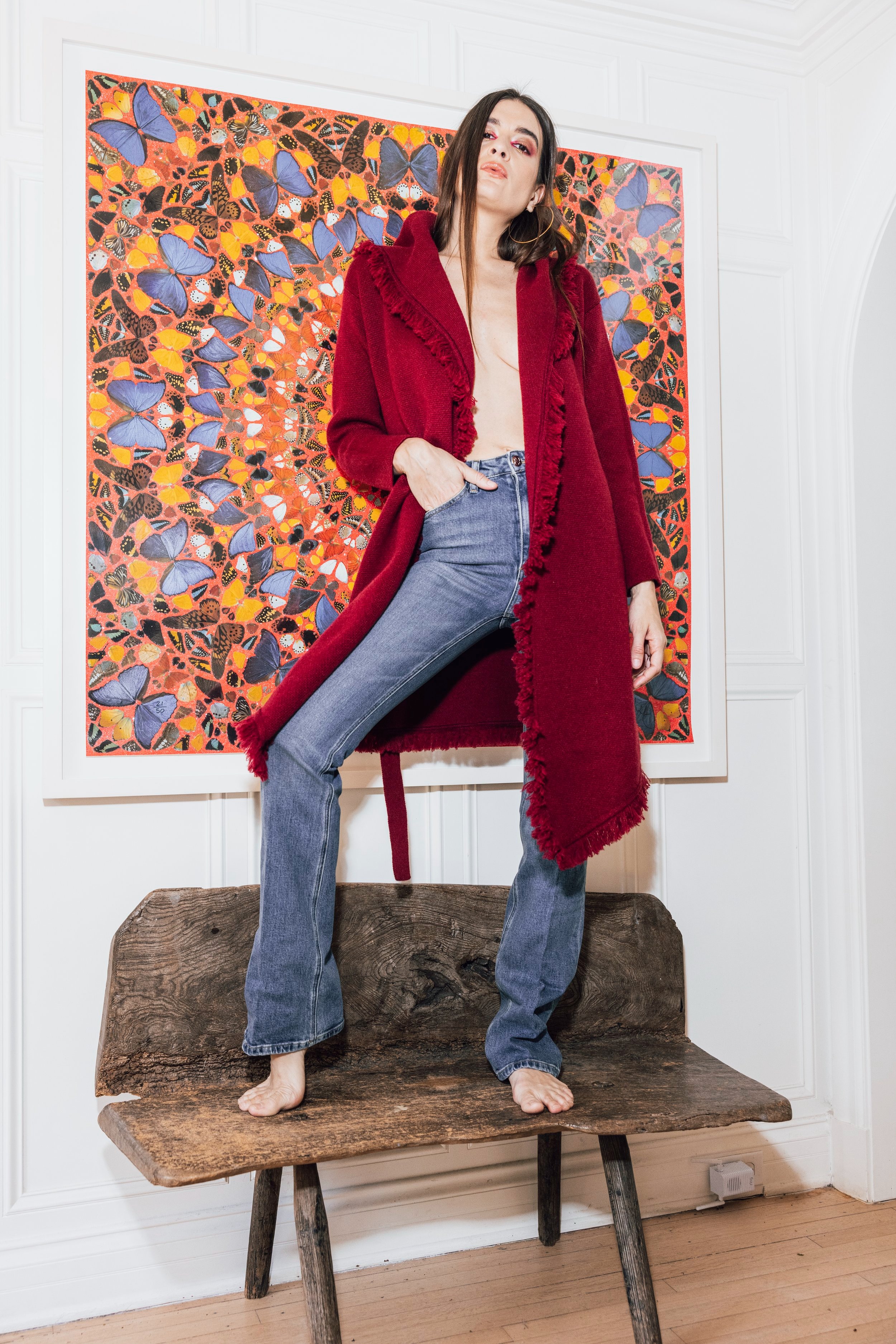 Full shot of a female model wearing a red fringe coat and jeans, standing barefoot on a wooden bench in front of an art print.