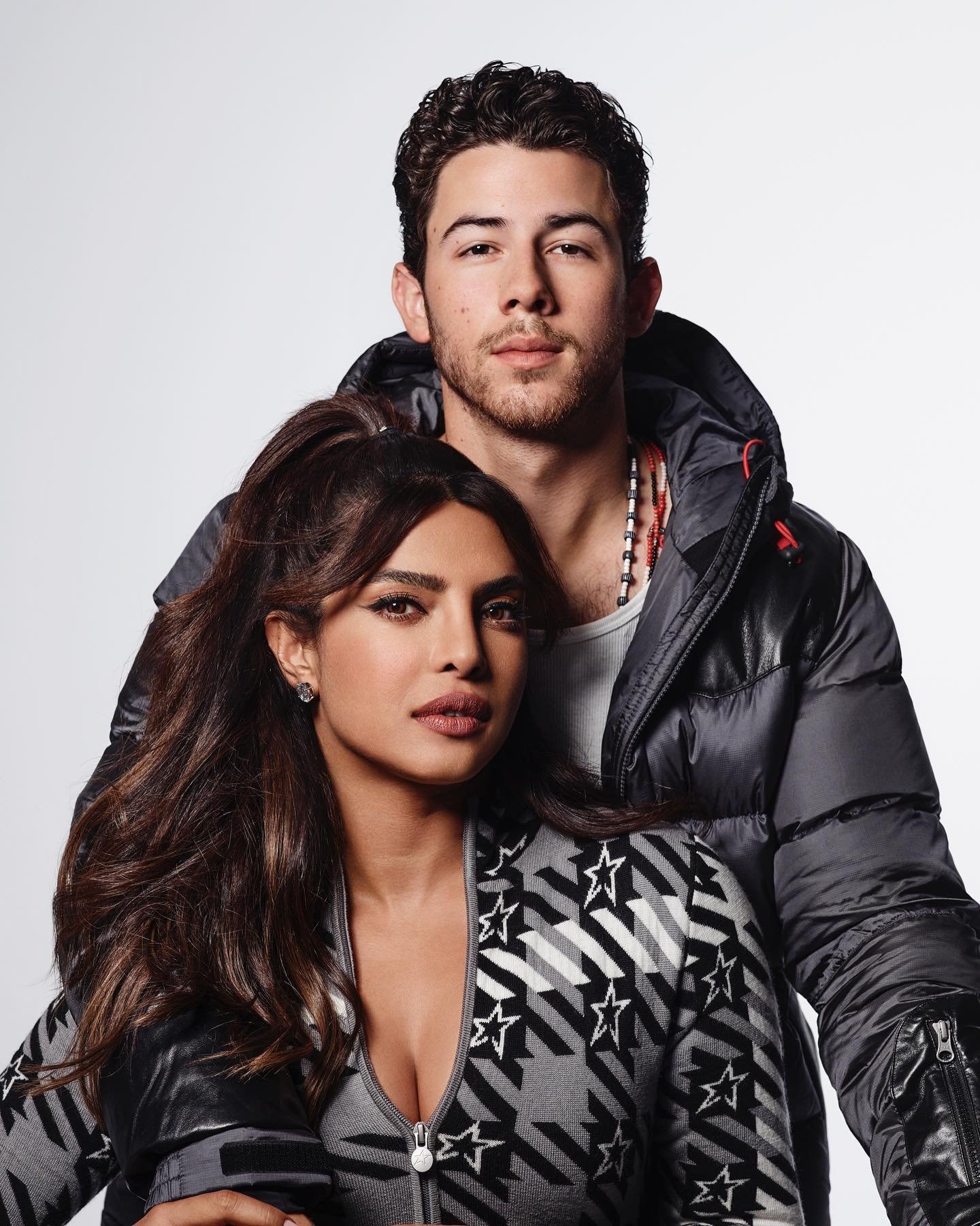 Studio shot featuring a man in a black puffer jacket standing behind a woman in a star-patterned jacket against a white backdrop.