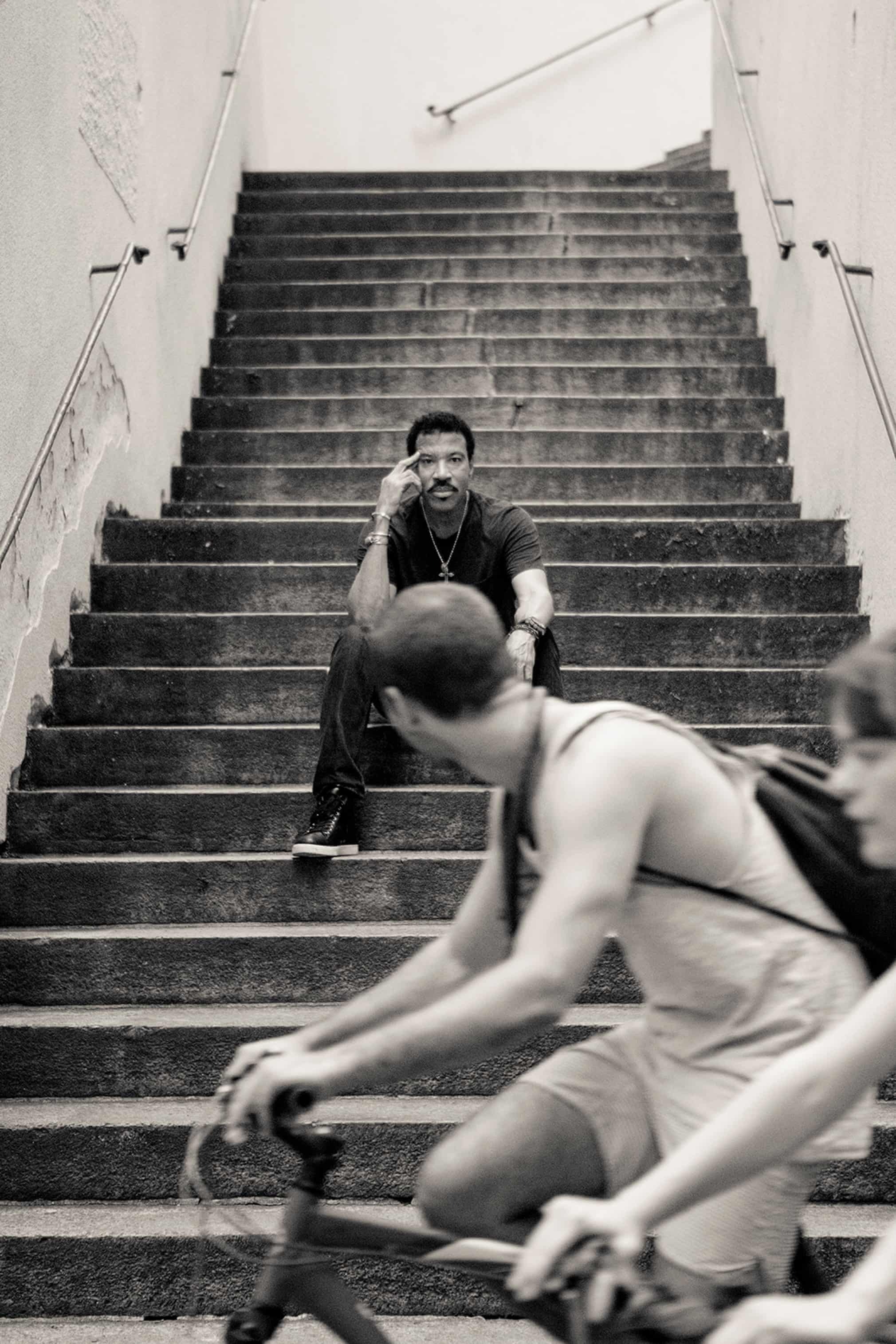 Black and white portrait of Lionel Richie sitting on concrete stairs looking pensively at the camera with cyclists in the foreground.