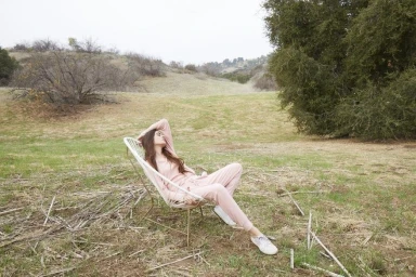 Woman in pink loungewear reclines in a white wire chair in a dry grassy meadow.