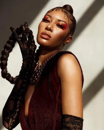 Close-up portrait of a stylish adult woman with braided hair, red eyeshadow, statement necklace, and a glittery burgundy dress, indoors.