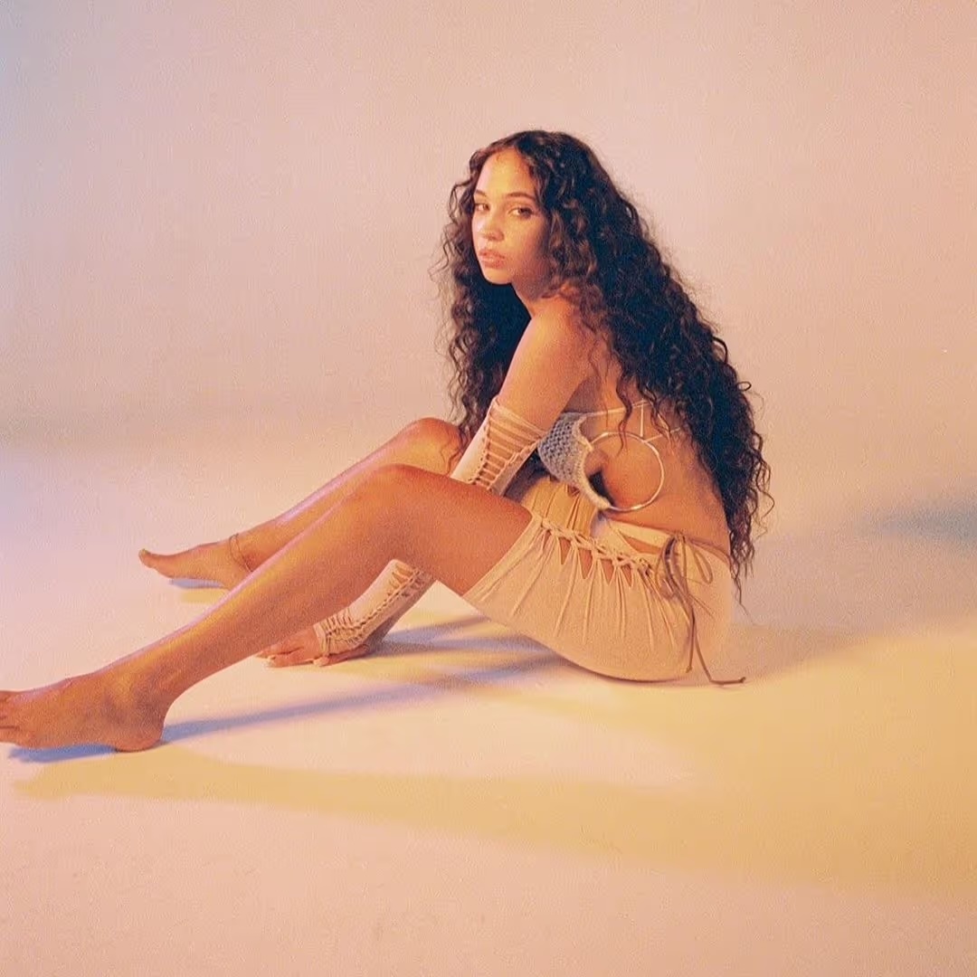 Young adult female model with long curly hair sits barefoot in a tan crocheted outfit in a brightly lit neutral studio setting.