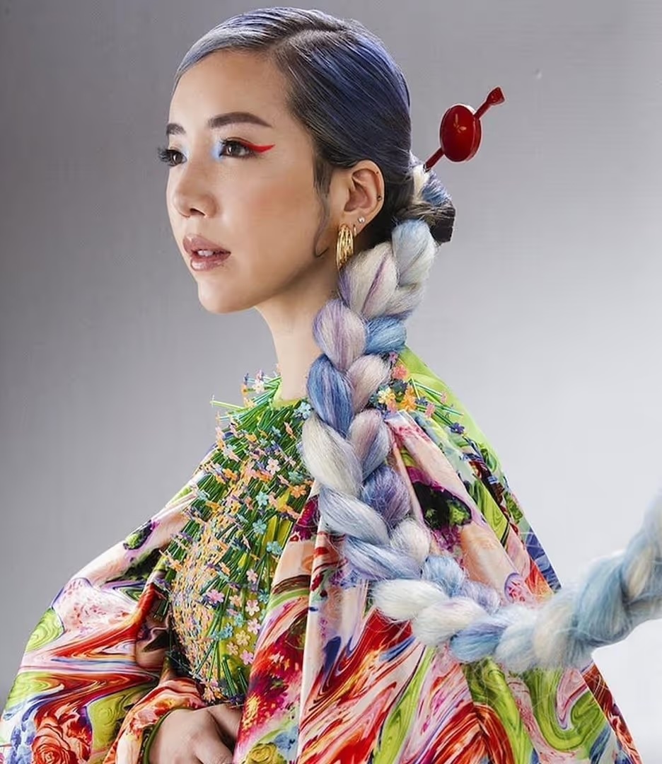 Adult female in a colorful patterned dress with a long braided hairstyle featuring a red hair stick, posing in front of a grey background.
