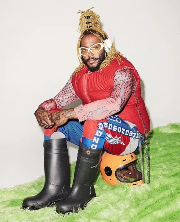 Adult male with beard, wearing white glasses, red vest, and black boots seated on a prop, set against white background.