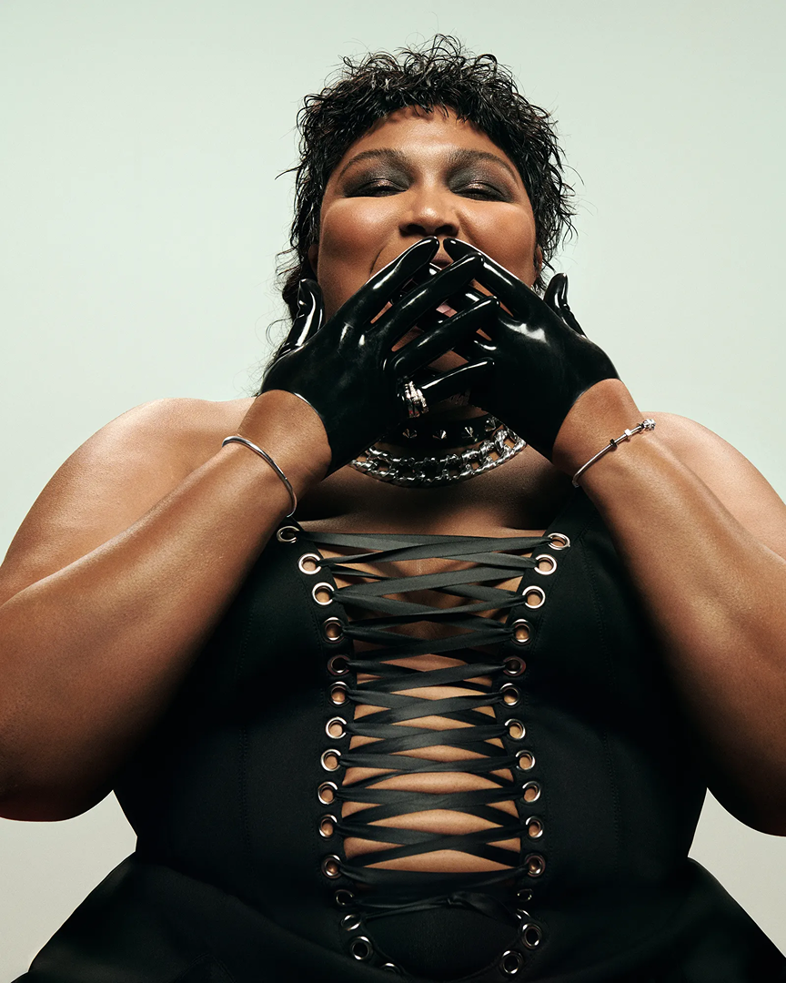 Close-up of Lizzo in black latex gloves and a black corset-style top with silver jewelry against a neutral background.