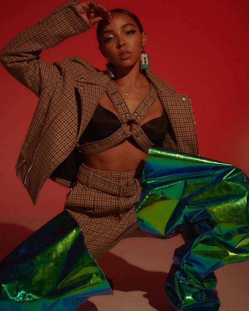 Young woman posing in plaid jacket and pants with a black top and iridescent chaps against a red backdrop in a studio shot.