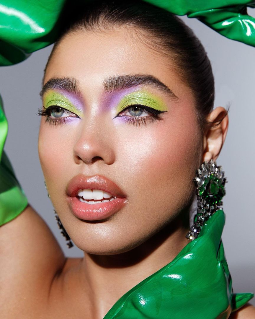 Close-up shot of a woman with green eye makeup, glossy pink lips, and large crystal earrings against a neutral grey background.