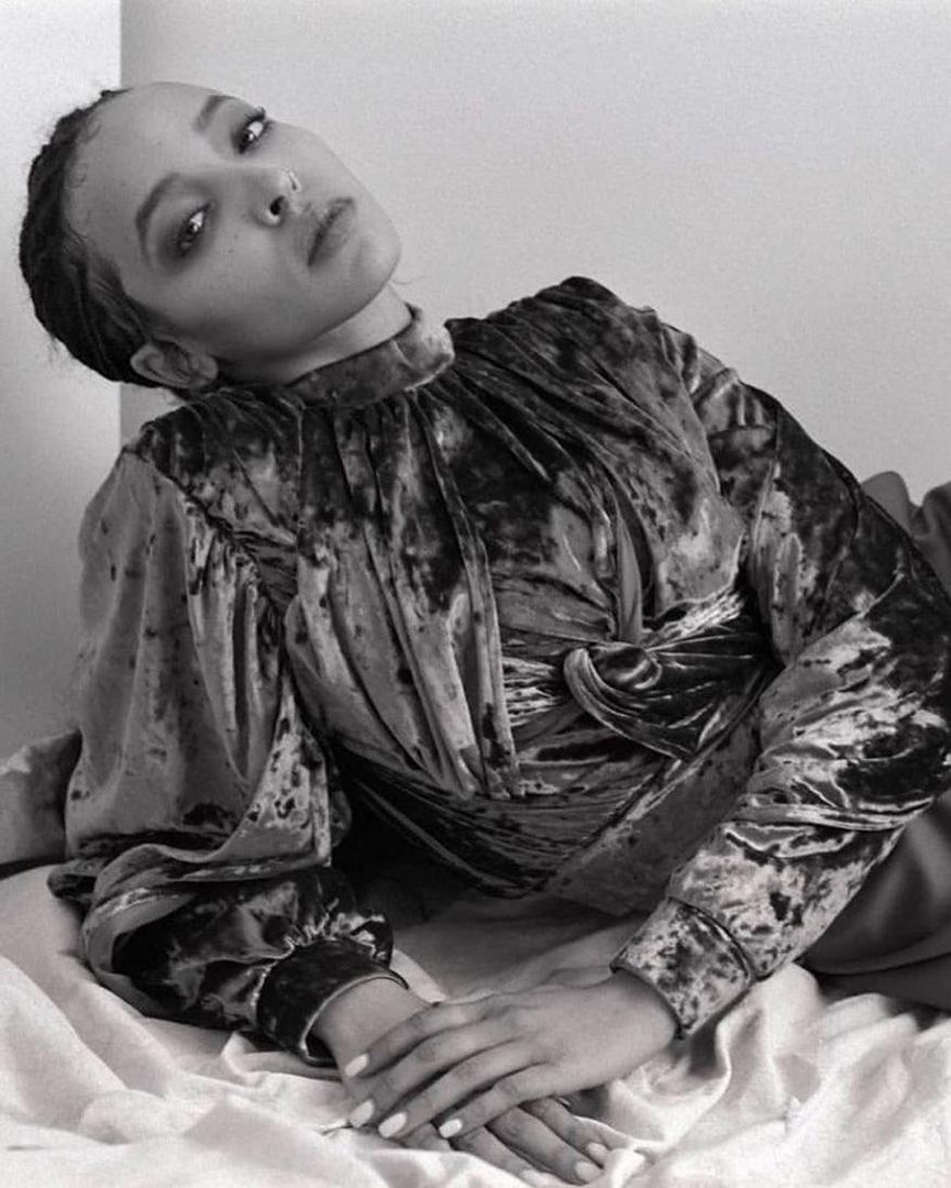 Black and white portrait of a woman in a velvet top lying on a bed with her hands together. The background is plain.