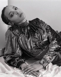 Portrait of a woman wearing a high-neck velvet top, posing on a bed with light bedding in a black and white composition.