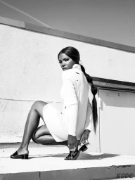 Black and white portrait of a woman crouching on a rooftop with dark hair in a long braided ponytail wearing a white top and sandals