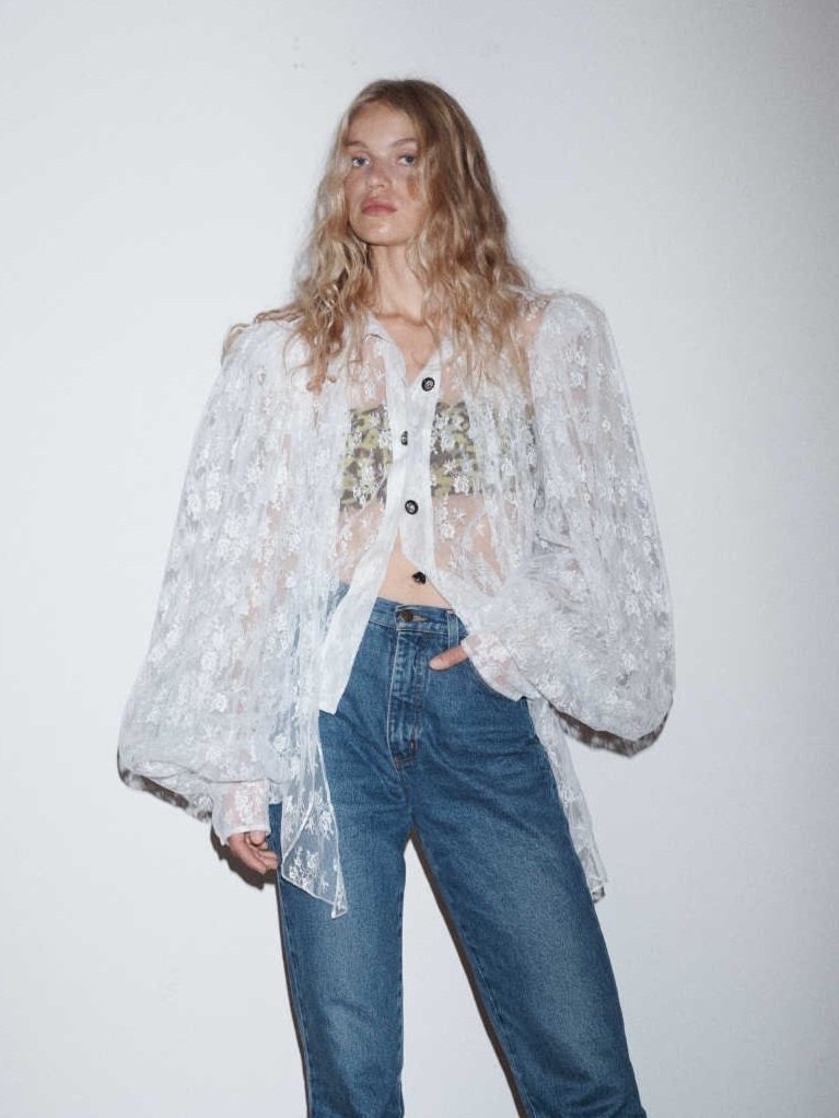 A woman with wavy blonde hair poses in a white lace shirt with blue jeans against a white wall, illuminated by soft lighting.