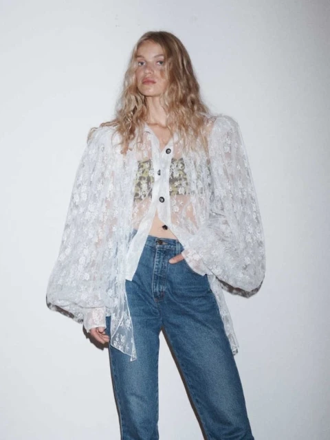 A woman with wavy blonde hair poses in a white lace shirt with blue jeans against a white wall, illuminated by soft lighting.