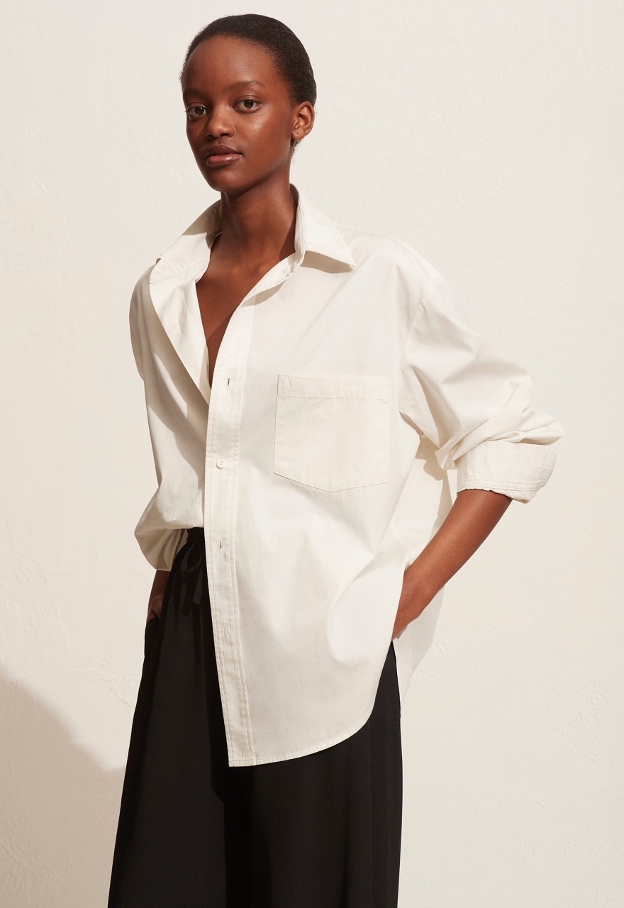 African American woman poses in an unbuttoned white shirt and black pants against an off-white background in a studio setting.