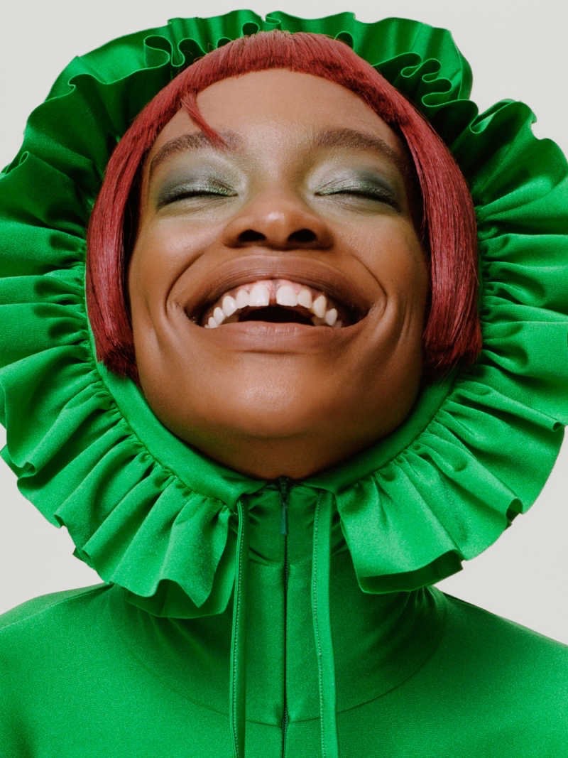 Close-up of a Black woman with short red hair, eyes closed, and a wide smile, wearing a green ruffled collar against a bright, neutral background.