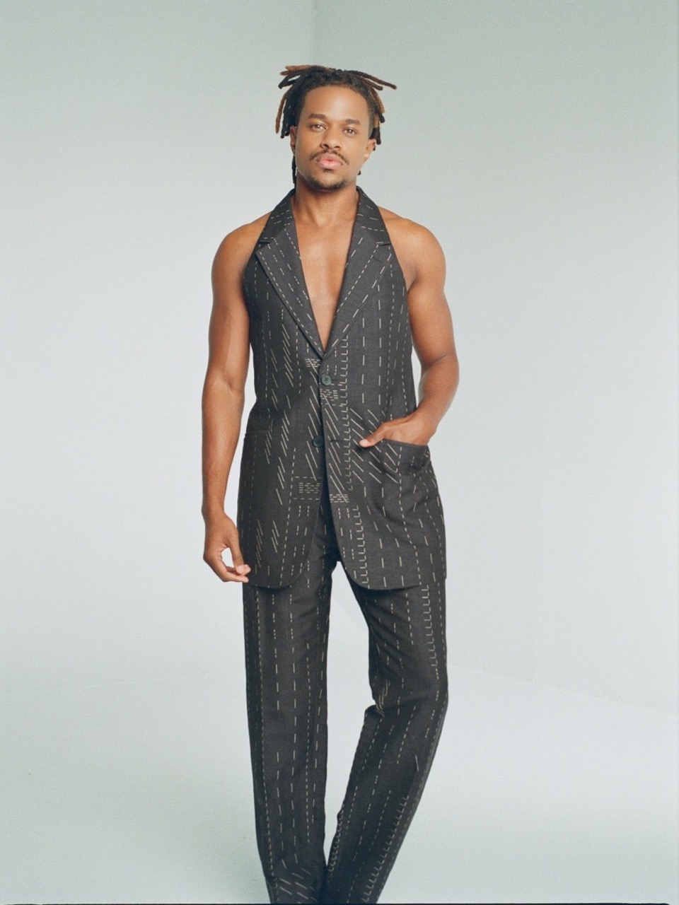 Adult male in a pinstripe sleeveless blazer and matching pants poses in a studio. He has dreadlocks and his hand is in his pocket.