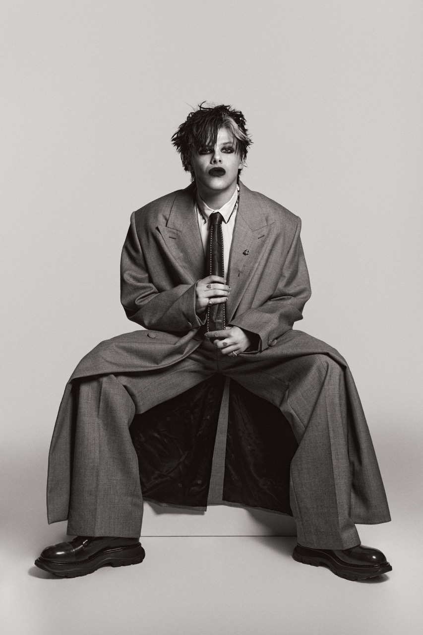 Portrait of a person with dark and white hair sitting in an oversized gray suit, holding a tie against a neutral background.