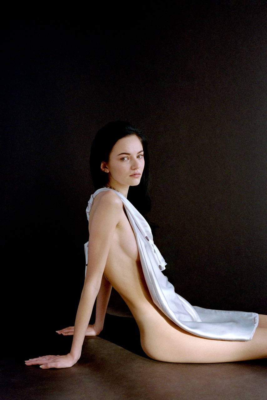 Elegant woman with dark hair poses against a black background, draped in a light cloth; studio lighting enhances her features.