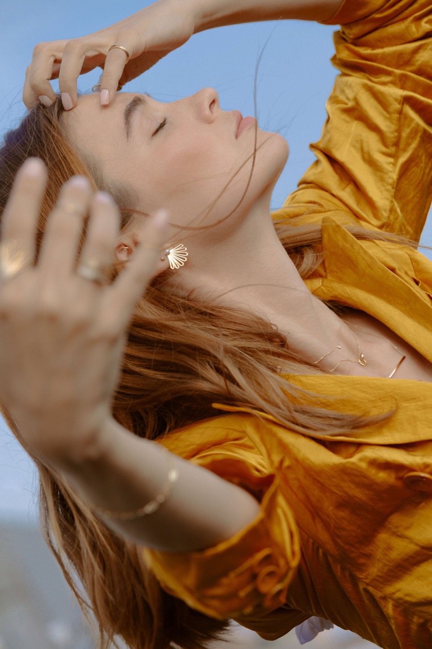 Woman in mustard-yellow jacket with hands near face, accessorized with seashell earrings, against blue sky backdrop in relaxed setting.