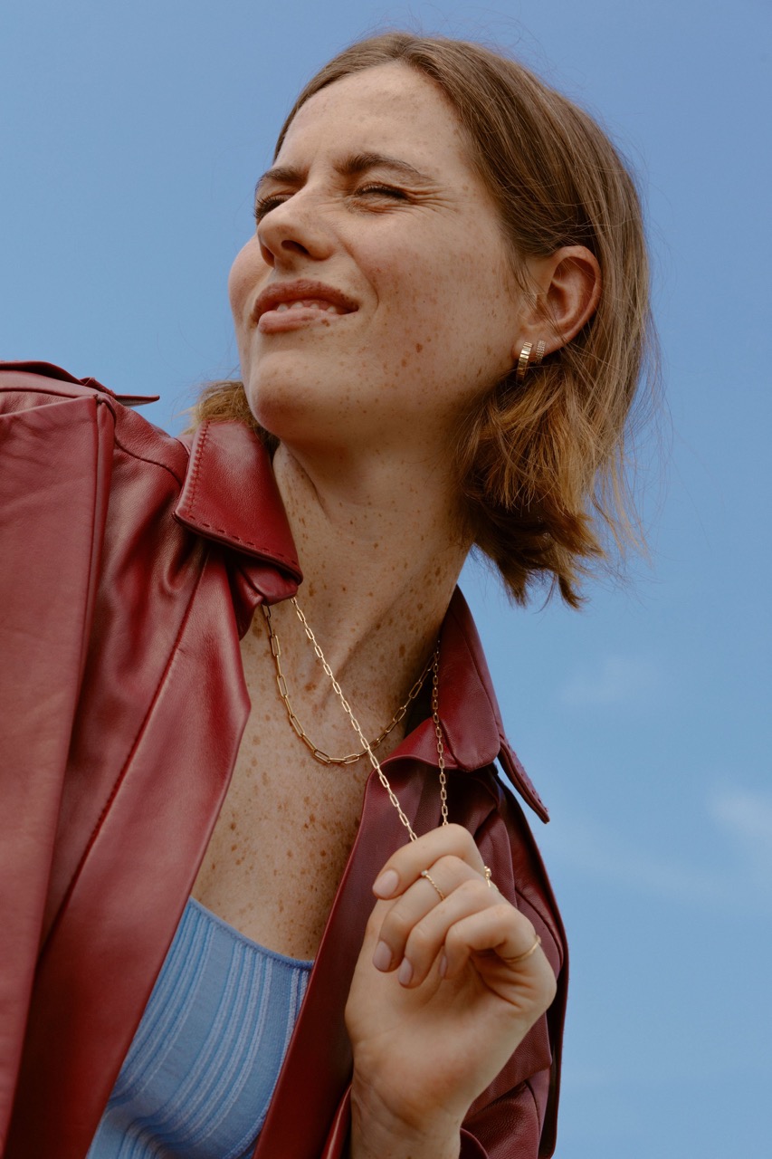 Woman in a red leather jacket against a bright blue sky. She has short auburn hair and is wearing gold jewelry.