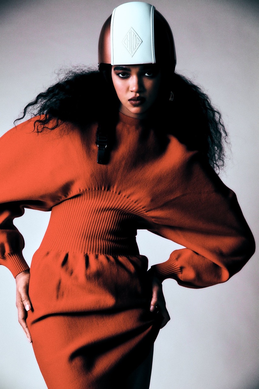 Woman with dark curly hair wearing a helmet and orange dress posing indoors in a studio, fashion shoot, studio lighting.