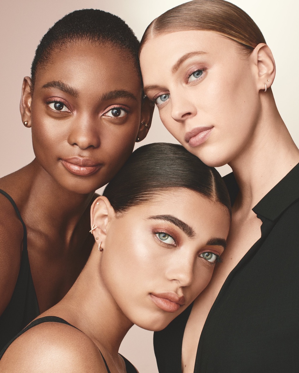 Three beautiful multi-ethnic women with clean makeup, posing together indoors against a neutral background in a close-up beauty shot.