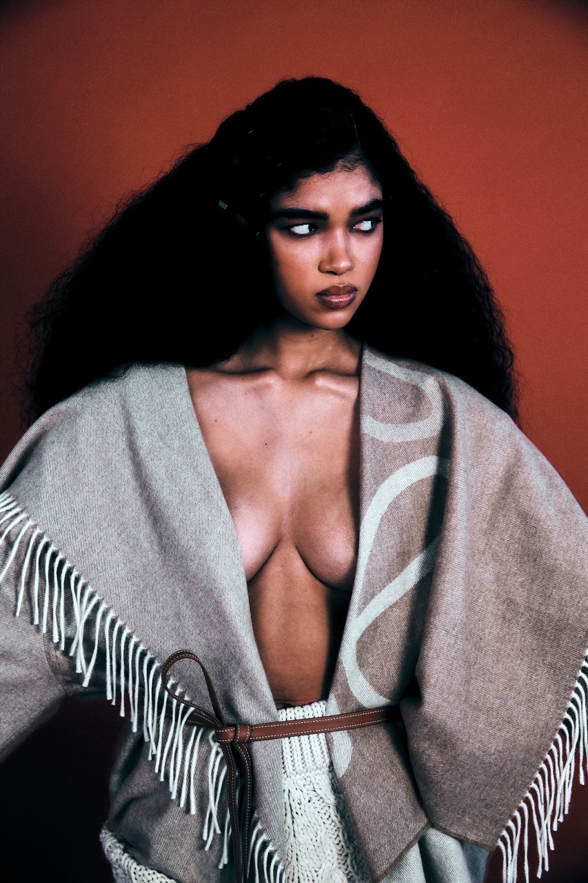 Portrait of woman with long black hair wearing a fringed poncho in front of an orange background in a fashion photography style image.