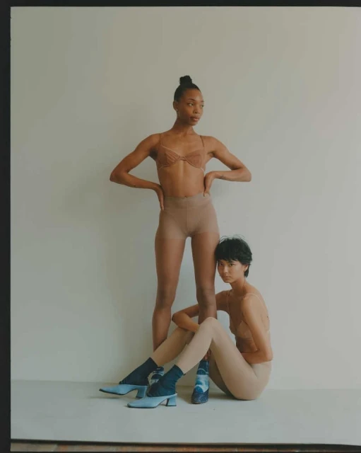 Two women pose indoors wearing lingerie and hosiery against a neutral wall, one standing and one seated with blue heeled shoes.