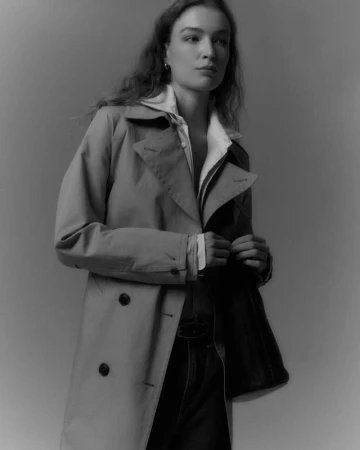 Woman wearing a beige trench coat over a white shirt against a plain dark studio background