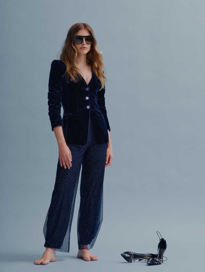 A barefoot woman in a dark velvet blazer and sequined pants poses in a studio, complemented by a pair of high heels nearby.