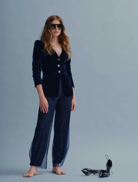 A barefoot woman in a dark velvet blazer and sequined pants poses in a studio, complemented by a pair of high heels nearby.