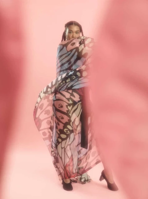 An adult female poses in a photo studio wearing a flowing butterfly-patterned garment and heeled shoes in front of a soft pink backdrop.