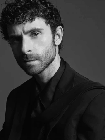 Portrait of a man with curly hair and beard wearing a dark suit against a grey studio background styled by Ali Kornhauser