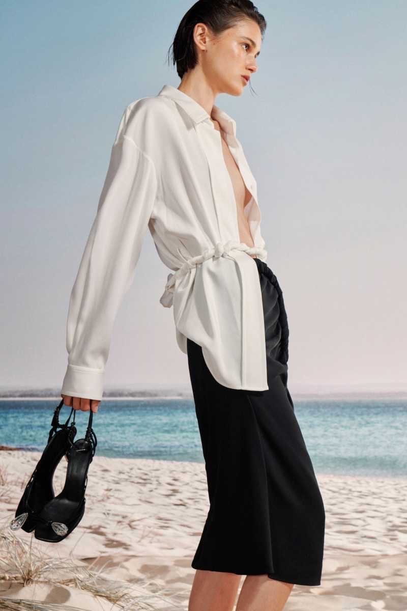 Woman on a beach wearing a white shirt and black skirt, holding black high heel shoes, with a blue ocean and sky background.